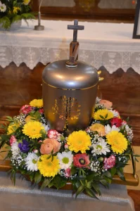 Urne aus gebürstetem Metall mit Holzkreuz, umgeben von einem Kranz aus bunten Frühlingsblumen