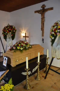 Aufbahrung mit hellem Sarg vor einem Kruzifix, Kränze und Kerzenständer