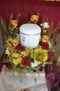 Weiße Urne mit Inschrift auf einem Kranz aus gelben und roten Rosen vor rotem Tuch