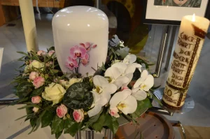 Weiße Urne mit Blumenschmuck aus Orchideen und Rosen, daneben eine bemalte Kerze