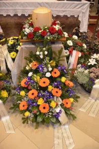 Urne mit Blumenschmuck in Orange und Violett, davor Kranzschleifen
