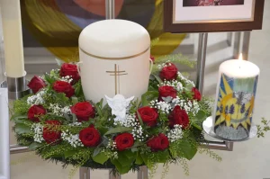 Weiße Urne mit Kreuz in einem Kranz aus roten Rosen, daneben eine verzierte Kerze