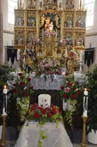 Kirchenraum mit goldenem Flügelaltar, davor ein Sarg mit Blumengirlanden