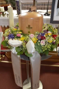 Helle Holzurne mit eingelegtem Kreuz auf einer Säule, davor bunter Blumenschmuck und Kerzen