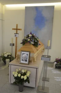 Aufbahrung mit hellem Holzsarg, Holzkreuz und Blumenschmuck in Violett und Weiß
