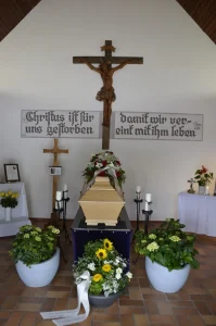 Aufbahrungsraum mit Kruzifix und Spruchtafel, Sarg und Blumenschmuck